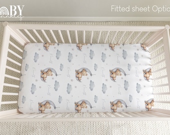 May include: A white fitted cot sheet with a repeating pattern of grey clouds, stars, and brown teddy bears holding onto a crescent moon. The sheet has the name "Knitt" printed on it.