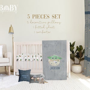 Op de afbeelding: Een 5-delige babybeddenset in grijs en wit met een groen alien-figuur. De set bevat een dekbed, een hoeslaken en drie decoratieve kussens. Het dekbed heeft een grijze achtergrond met witte sterren en een groen alien-figuur met de naam "JACKSON" eronder. Een van de decoratieve kussens heeft de tekst "WHEREVER I GO, HE GOES".
