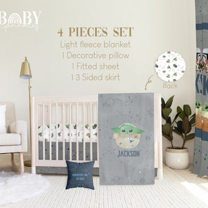 Op de afbeelding: Een vierdelige babybeddenset met een grijs en wit ruimtethema met een groen alienfiguur. De set bevat een licht fleece deken, een decoratief kussen, een hoeslaken en een 13-zijdige rok. Het kussen heeft de tekst "WHEREVER I GO HE GOES" erop gedrukt. De rok heeft de naam "JACKSON" erop gedrukt.