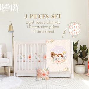 May include: A 3-piece baby bedding set with a light pink and floral design. The set includes a fitted sheet, a decorative pillow, and a fleece blanket with a personalized name and a floral wreath. The blanket has a pink and white floral pattern on the back.