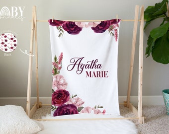 Può includere: Una copertina per neonati bianca con un motivo floreale in tonalità rosa e bordeaux. La copertina ha il nome "Agatha Marie" scritto in corsivo.