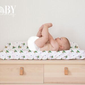 May include: A white changing pad cover with a repeating pattern of green and brown baby Yoda characters and pink and blue stars. A baby is lying on the changing pad cover.