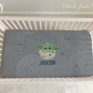 Op de afbeelding: Grijze getailleerde wieg lakens met een ruimtethema met een groene alien, planeten en sterren. De lakens hebben de naam "JACKSON" erop gedrukt.