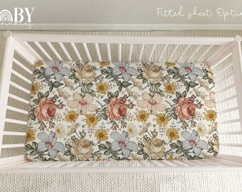 Puede incluir: Una cuna blanca con una sábana ajustable con un patrón floral en tonos amarillos, rojos y azules. La sábana tiene un fondo blanco con una variedad de flores y hojas. El texto "Fitted sheet Option |" es visible en la esquina superior derecha de la imagen.