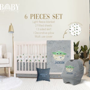 Op de afbeelding: Een 6-delige babybeddenset met een grijs en wit ruimtethema met een groen alien-personage. De set bevat een licht fleece deken, twee hoeslakens, een 3-zijdige rok, een decoratief kussen en een multifunctionele hoes. De set is gepersonaliseerd met de naam "JACKSON".