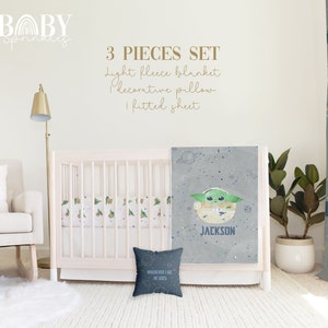 Op de afbeelding: Een driedelige babybeddenset met een grijs en wit hoeslaken, een grijs fleece deken met een groen alienfiguur en de naam "JACKSON" erop gedrukt, en een grijs decoratief kussen met de tekst "WHEREVER I GO HE GOES".