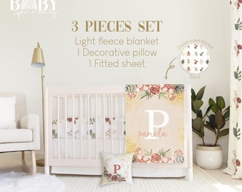 Puede incluir: Un juego de ropa de cama para bebé de 3 piezas con un diseño floral. El juego incluye una manta de microfibra ligera, una almohada decorativa y una sábana ajustable. La manta presenta un patrón floral con un fondo de brillo dorado y el nombre "pamela" escrito en blanco. La almohada es de forma cuadrada con un diseño geométrico y la letra "P" en el centro. La sábana ajustable tiene un patrón floral sobre un fondo blanco.