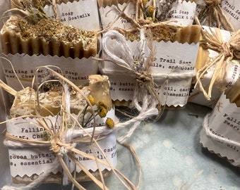 May include: A collection of handmade soap bars wrapped in brown paper and twine. The soap bars are labeled with names like "Falling Star", "Trail Soap", and "Pear, Cinnamon".