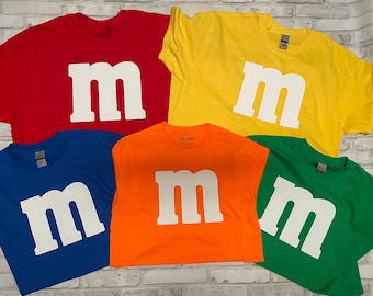 May include: Five t-shirts with a white "M" printed on them. The shirts are red, yellow, orange, blue, and green.