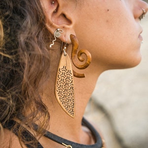 May include: A woman wearing a pair of wooden earrings. One earring is a long, thin, leaf-shaped piece with a geometric pattern carved into it. The other earring is a shorter, thicker, spiral-shaped piece.