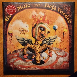 May include: A vinyl record album cover with a colorful illustration of a voodoo doll with wings, flames, and a pincushion. The album title is "Mule ∞ Déjà Voodoo" and the text "Coloured Vinyl Limited Edition" is in a circle on the top left corner.