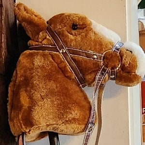May include: A brown plush horse head with a white patch on its forehead. The horse is wearing a brown leather bridle with a white stitching detail and a brown leather strap with a silver buckle. The horse's head is mounted on a wooden stick.
