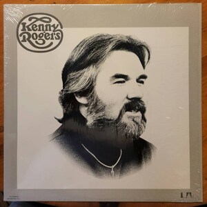 May include: A black and white photo of Kenny Rogers on a vinyl record album cover. The album cover is gray with a white border. The album title is not visible.