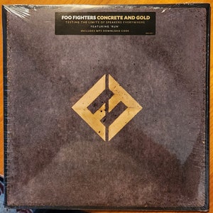 May include: A vinyl record album cover for Foo Fighters' Concrete and Gold album. The cover features a textured, dark gray background with a gold diamond-shaped logo in the center. The album title, "Concrete and Gold", is printed in white text at the top of the cover. The text "Testing the limits of speakers everywhere" is printed in white text below the album title. The text "Featuring 'Run'" is printed in white text below the previous text. The text "Includes MP3 download code" is printed in white text below the previous text.