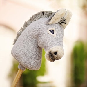 Pu&ograve; includere: Un giocattolo di peluche grigio a forma di asino con un manico di legno. L'asino ha una faccia bianca con occhi neri e un naso nero.