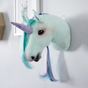 Pu&ograve; includere: Una testa di unicorno blu chiaro con un corno viola e una criniera turchese e viola. La testa di unicorno &egrave; montata su una parete bianca.