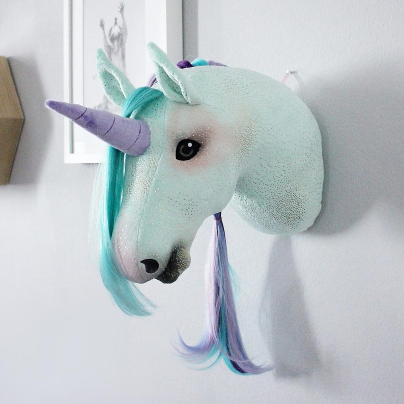 Pu&ograve; includere: Una testa di unicorno blu chiaro con un corno viola e una criniera turchese e viola. La testa di unicorno &egrave; montata su una parete bianca.