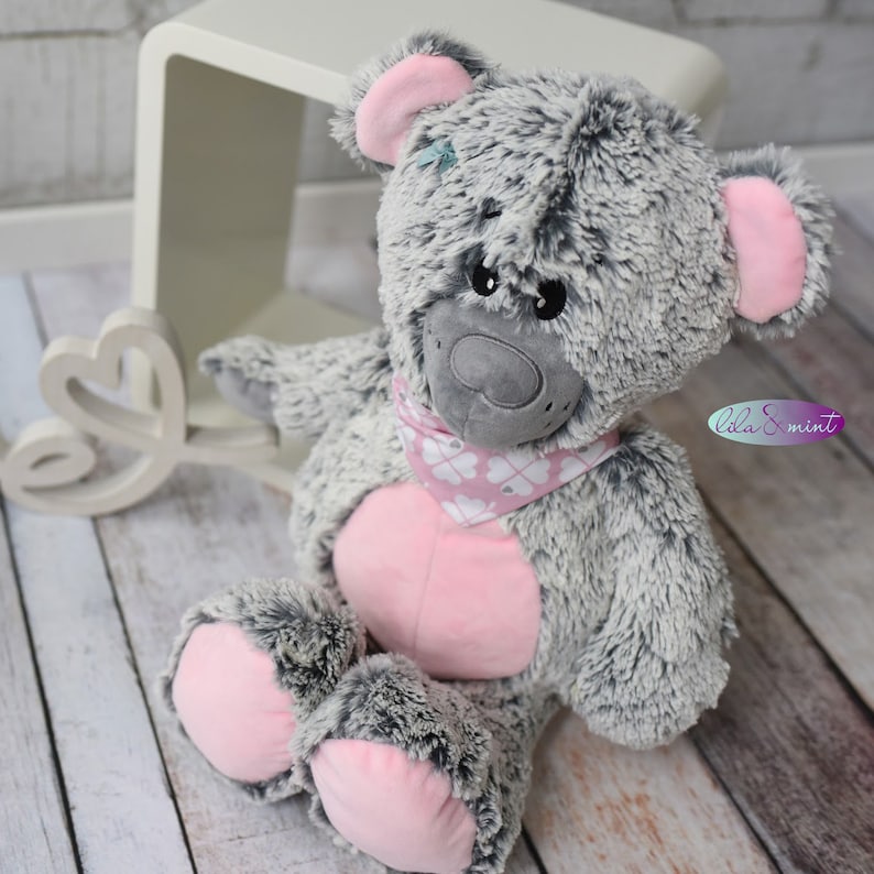 Puede incluir: Un oso de peluche gris y rosa con una pajarita rosa. El oso tiene una nariz gris y ojos negros. El oso est&aacute; sentado sobre una superficie de madera.