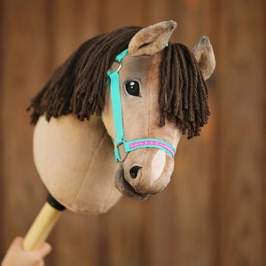 Pu&ograve; includere: Una testa di cavallo di peluche marrone e beige con un morsetto turchese e rosa. Il cavallo ha una lunga criniera e coda marroni.