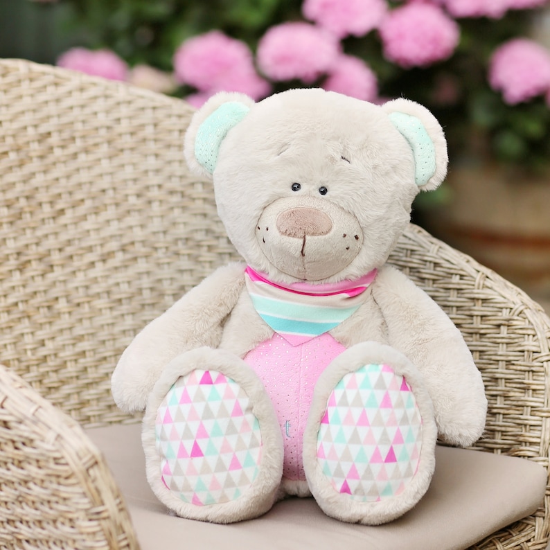 Puede incluir: Un oso de peluche beige con detalles en rosa y azul se sienta en una silla de mimbre. El oso lleva una bufanda a rayas rosa y azul y pies con estampado rosa y blanco.