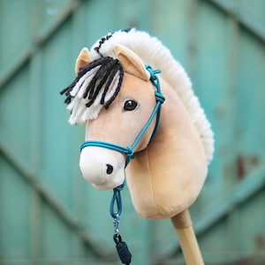 Pu&ograve; includere: Una testa di cavallo di peluche con una criniera bianca e nera e un guinzaglio di corda blu turchese. Il cavallo &egrave; beige e ha il naso marrone.
