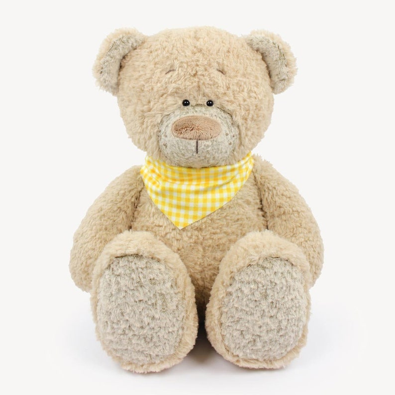 Puede incluir: Un oso de peluche con una bandana a cuadros amarillos y blancos. El oso est&aacute; sentado con las piernas cruzadas y tiene un pelaje suave y marr&oacute;n.