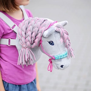 Pu&ograve; includere: Una testa di cavallo di peluche grigia con una criniera intrecciata rosa e grigia e un morsetto blu e turchese. Il cavallo &egrave; attaccato a un'imbracatura bianca indossata da un bambino.