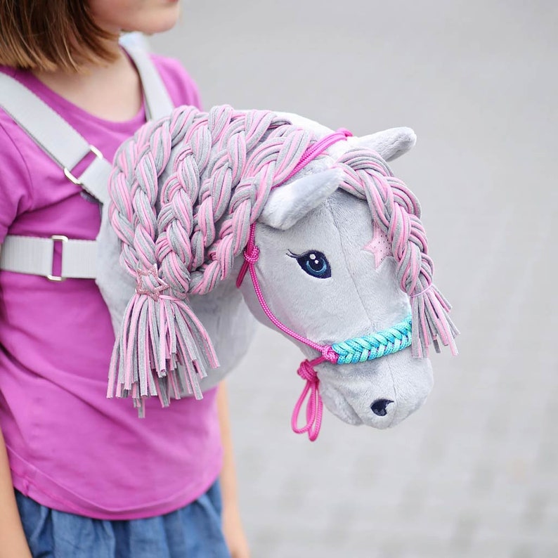 Pu&ograve; includere: Una testa di cavallo di peluche grigia con una criniera intrecciata rosa e grigia e un morsetto blu e turchese. Il cavallo &egrave; attaccato a un'imbracatura bianca indossata da un bambino.