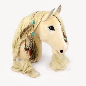 Pu&ograve; includere: Una testa di cavallo di peluche beige con una lunga criniera e coda bionde. Il cavallo indossa una fascia intrecciata con piume e perline turchesi e rosse.