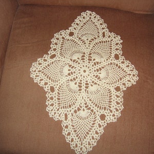 May include: A white crocheted doily with a delicate, intricate design. The doily is laid out on a brown fabric surface.