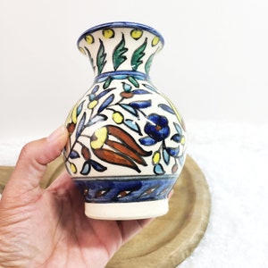 May include: A small, white ceramic vase with a blue and yellow floral pattern. The vase has a round body and a narrow neck.