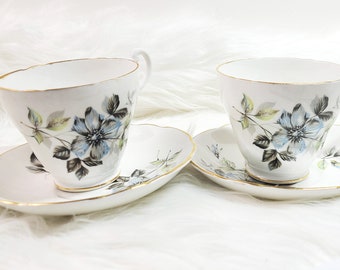May include: Two white teacups and saucers with a floral pattern. The cups and saucers have a gold trim. The flowers are blue, green, and gray.