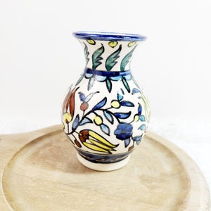May include: A small, hand-painted ceramic vase with a white base and a blue rim. The vase features a colorful floral design with blue, yellow, red, and green accents. The vase is displayed on a light brown wooden tray.