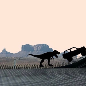 May include: A black silhouette of a Tyrannosaurus Rex dinosaur chasing a black silhouette of a jeep against a desert landscape background.