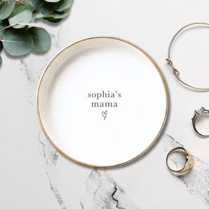 May include: A round white ceramic trinket dish with a gold-colored rim. The center of the dish features the black text "sophia's mama" above a small hand-drawn heart symbol. The dish is surrounded by a gold bangle, a ring, and a band.