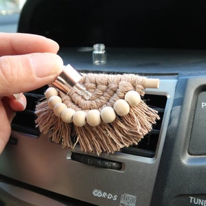 Può includere: Un diffusore per auto in macramé marrone e beige con perline di legno e una piccola bottiglia di vetro con tappo color oro rosa. Il diffusore è appeso a una bocchetta di ventilazione dell'auto.