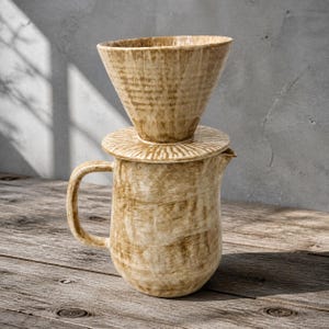 May include: A ceramic coffee pour-over dripper set featuring a conical filter holder resting on a matching pitcher with a handle. The set has a textured, mottled tan and beige glaze with visible brushstroke patterns and a radial ribbed lid.
