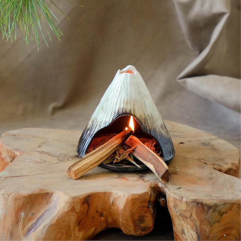 May include: A ceramic volcano-shaped incense burner with a lit flame inside. The burner is white and black, with a textured surface. Two pieces of wood are placed inside the burner. The burner sits on a wooden surface.