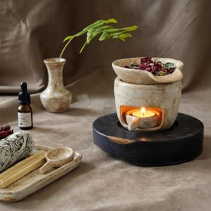 Op de afbeelding: Een crèmekleurige keramische etherische olieverspreider met een brandende kaars erin, op een zwarte houten voet. De diffuser heeft een bovenste kom gevuld met gedroogde bloemen. Een kleine vaas en een oliefles staan op de achtergrond.