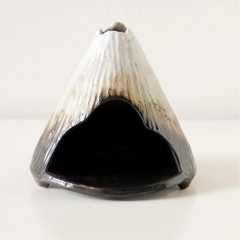 May include: A ceramic triangular sculpture with a mountain-like shape. The top is white, transitioning to brown and black at the base. It features a large, open, black interior space. The sculpture is on a white surface.