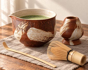 Puede incluir: Un cuenco de cerámica para matcha con pico vertedor que contiene té verde, un soporte de cerámica a juego para el batidor y una cuchara de té de bambú. El cuenco y el soporte presentan un esmalte marrón oscuro con patrones de pinceladas blancas.