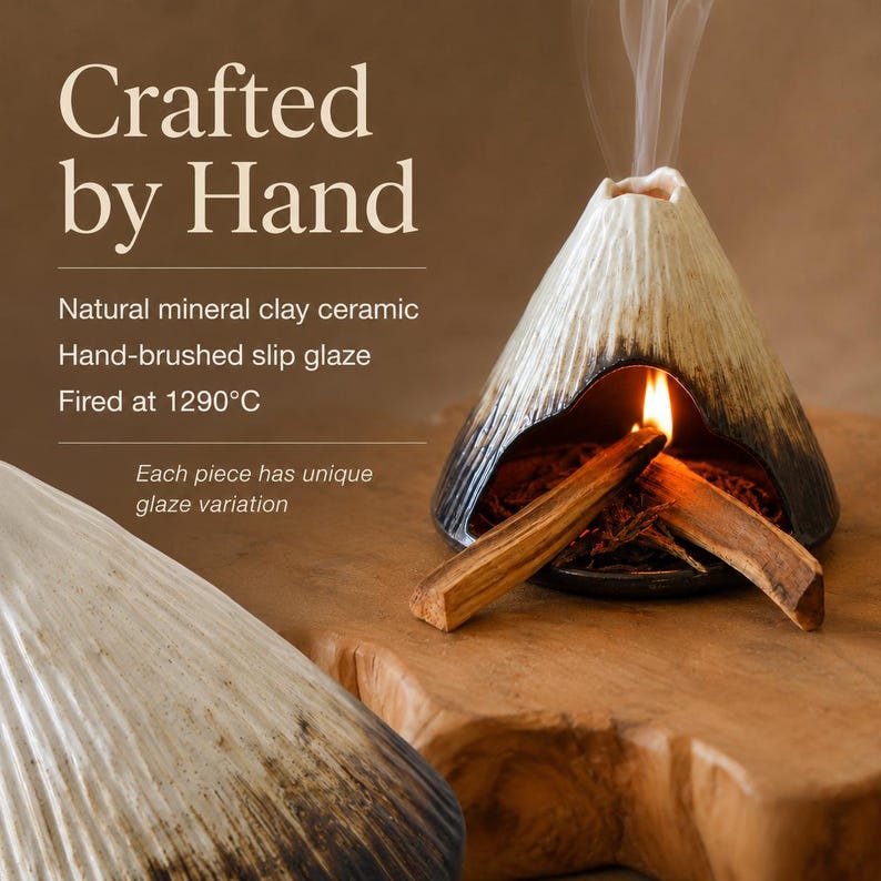 May include: A ceramic incense holder in the shape of a volcano, with a light-colored exterior and dark accents. The text "Crafted by Hand" is at the top. The holder is lit with a flame and has two wooden sticks inside.