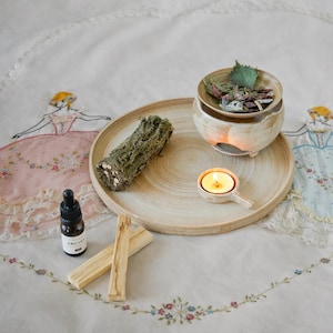 Op de afbeelding: Een serene opstelling met een houten dienblad met een keramische olie diffuser met gedroogde kruiden, een brandende kaars, een bundel gedroogde schors, palo santo stokken en een klein flesje etherische olie. De items staan op een bloemen geborduurd doek.