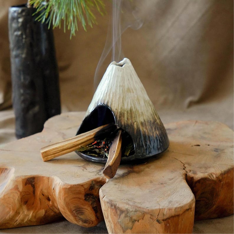 May include: A ceramic incense burner shaped like a volcano, with a white and brown gradient. Incense sticks are placed inside, emitting smoke. The burner sits on a wooden slab, with a dark vase in the background.