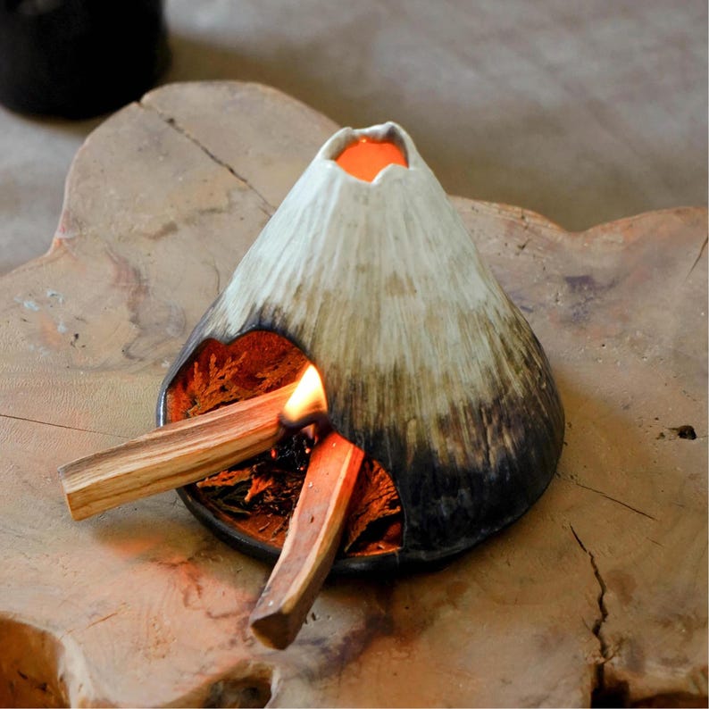 May include: A ceramic volcano-shaped incense holder with a white and brown exterior. Two pieces of burning palo santo wood are placed in the holder, with flames visible. The holder sits on a wooden surface.