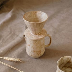May include: A beige ceramic coffee pour-over set. It features a cone-shaped dripper, a circular base, and a small jug with a handle. The textured surface gives a rustic, handmade appearance. The set is designed for brewing coffee, ideal for home use.