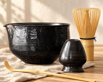 Può includere: Un set da tè matcha composto da una grande ciotola in ceramica nera testurizzata con beccuccio, un supporto per frusta in ceramica nera, una frusta in bambù chiaro con setole sottili e un cucchiaio da tè in bambù lungo, sottile e curvo.