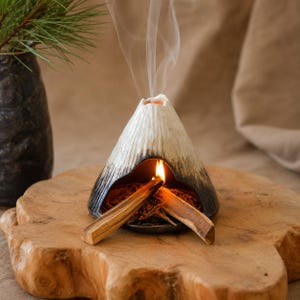 May include: A ceramic incense holder shaped like a volcano, with a black base and white and gray textured sides. Burning incense emits smoke. Two pieces of wood are lit inside the holder, resting on a wooden slab.