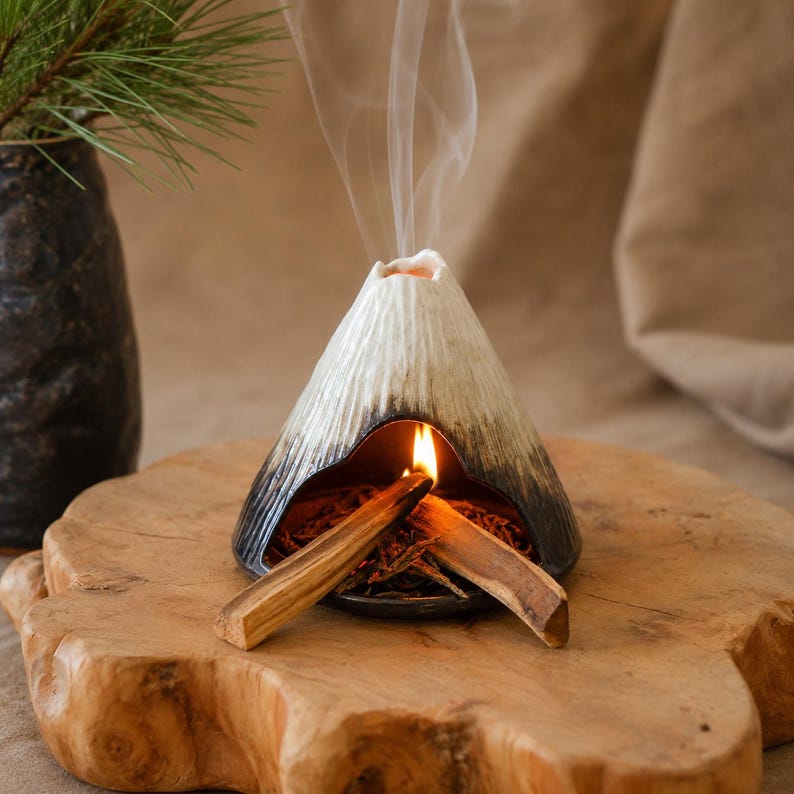 May include: A ceramic incense holder shaped like a volcano, with a black base and white and gray textured sides. Burning incense emits smoke. Two pieces of wood are lit inside the holder, resting on a wooden slab.