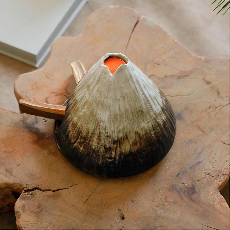 May include: A ceramic volcano-shaped decorative object with a dark brown base, transitioning to a lighter, textured surface, and an orange interior. The object is placed on a wooden surface, showcasing its unique design and craftsmanship.